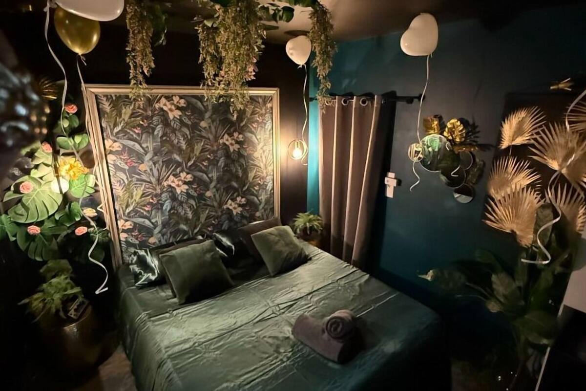 A bedroom with a bed, green bedding, a floral wall mural, and various potted plants.