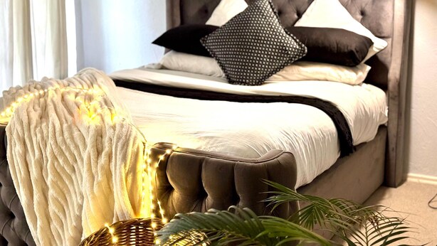 A bedroom with a tufted headboard, a bed with white bedding, two black pillows, and a bedside lamp. - Duke Mews Loft: Cozy 1-Bedroom Apartment in Sandton: 15 Mins to Sandton City. (Sandton)