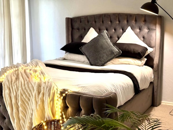 Une chambre à coucher avec une tête de lit capitonnée, un lit avec une literie blanche, deux oreillers noirs et une lampe de chevet.
