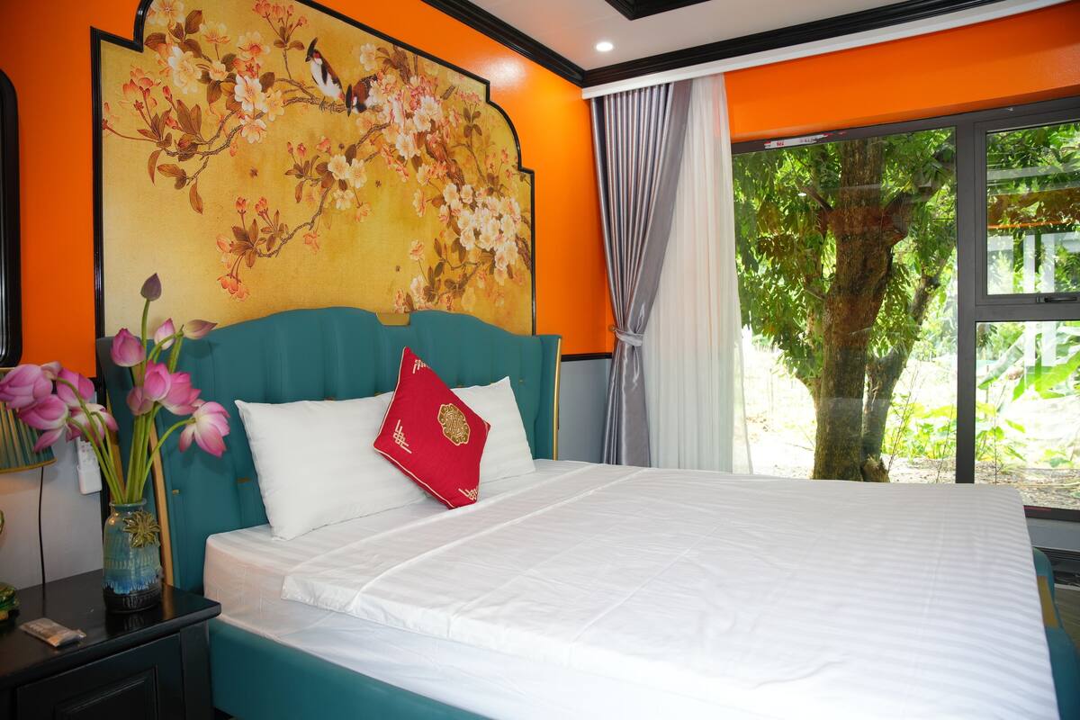 Un dormitorio con una cama grande, un mural floral en la pared, una ventana con vistas a un árbol y un florero con flores en la mesita de noche.
