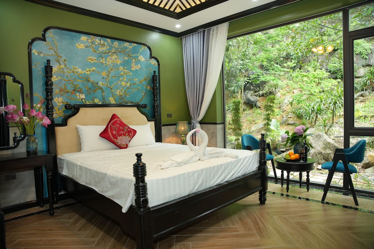 Un dormitorio con una cama grande, cabecera con diseño floral, almohada roja, mesita de noche con lámpara y vistas a la vegetación exterior.