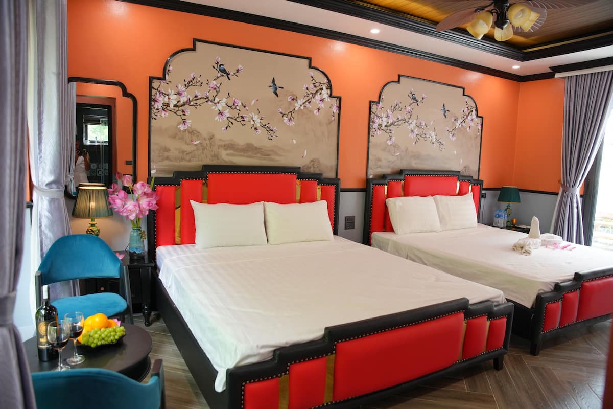 Habitación de hotel con dos camas, cada una con cabeceros tapizados en rojo y ropa de cama blanca. Hay una mesita con una lámpara y un florero con flores.