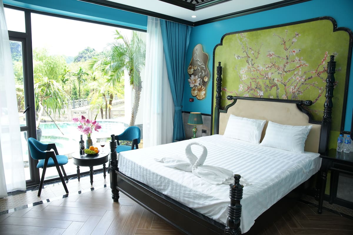 Habitación de hotel con una cama grande, una mesita con un florero y fruta, una silla y vistas a una piscina y zonas verdes.