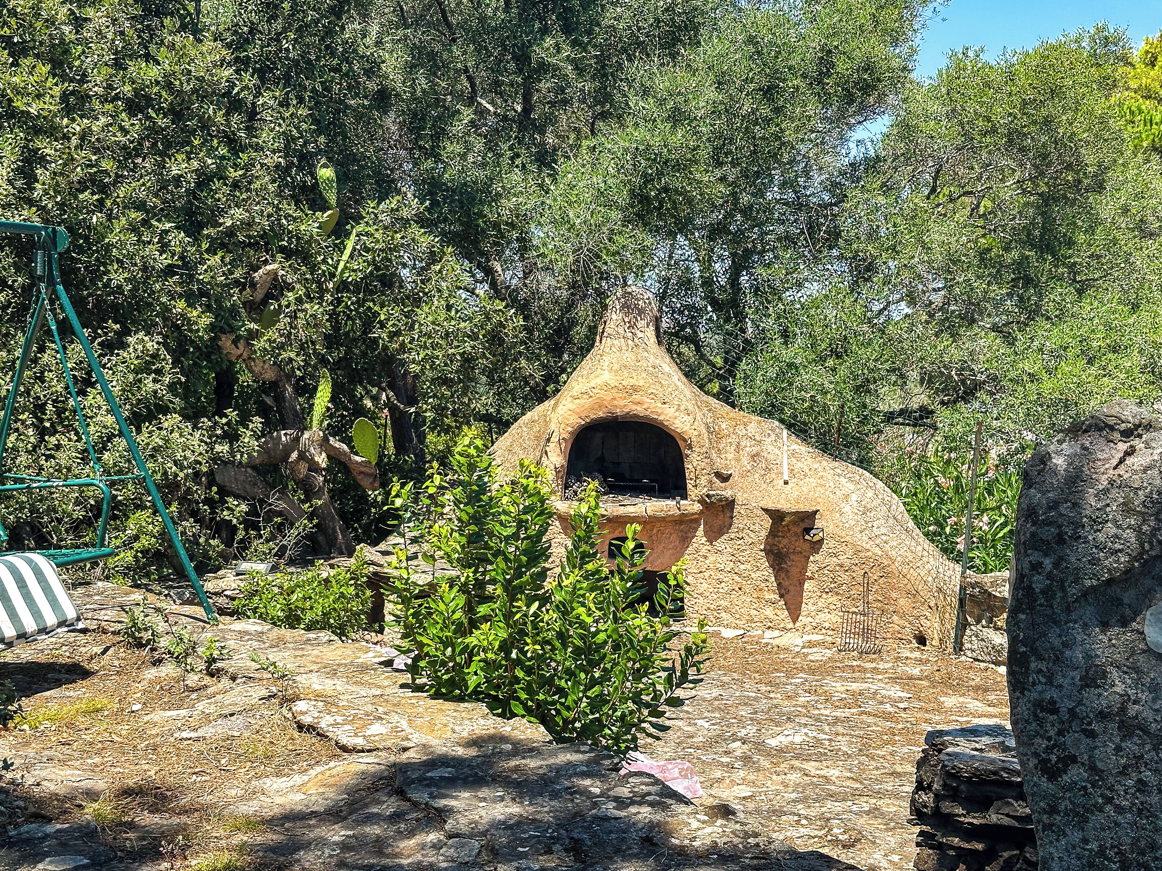 Een rustieke omgeving buiten, met een stenen structuur die op een hut lijkt, omgeven door groen en bomen.