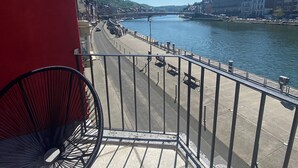 Un balcón con una silla de metal, con vistas a un paseo fluvial con edificios y un puente al fondo.