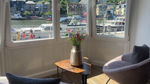 Una habitación con un amplio ventanal con vistas a un puerto deportivo, una mesita con un jarrón de flores y dos sillas.