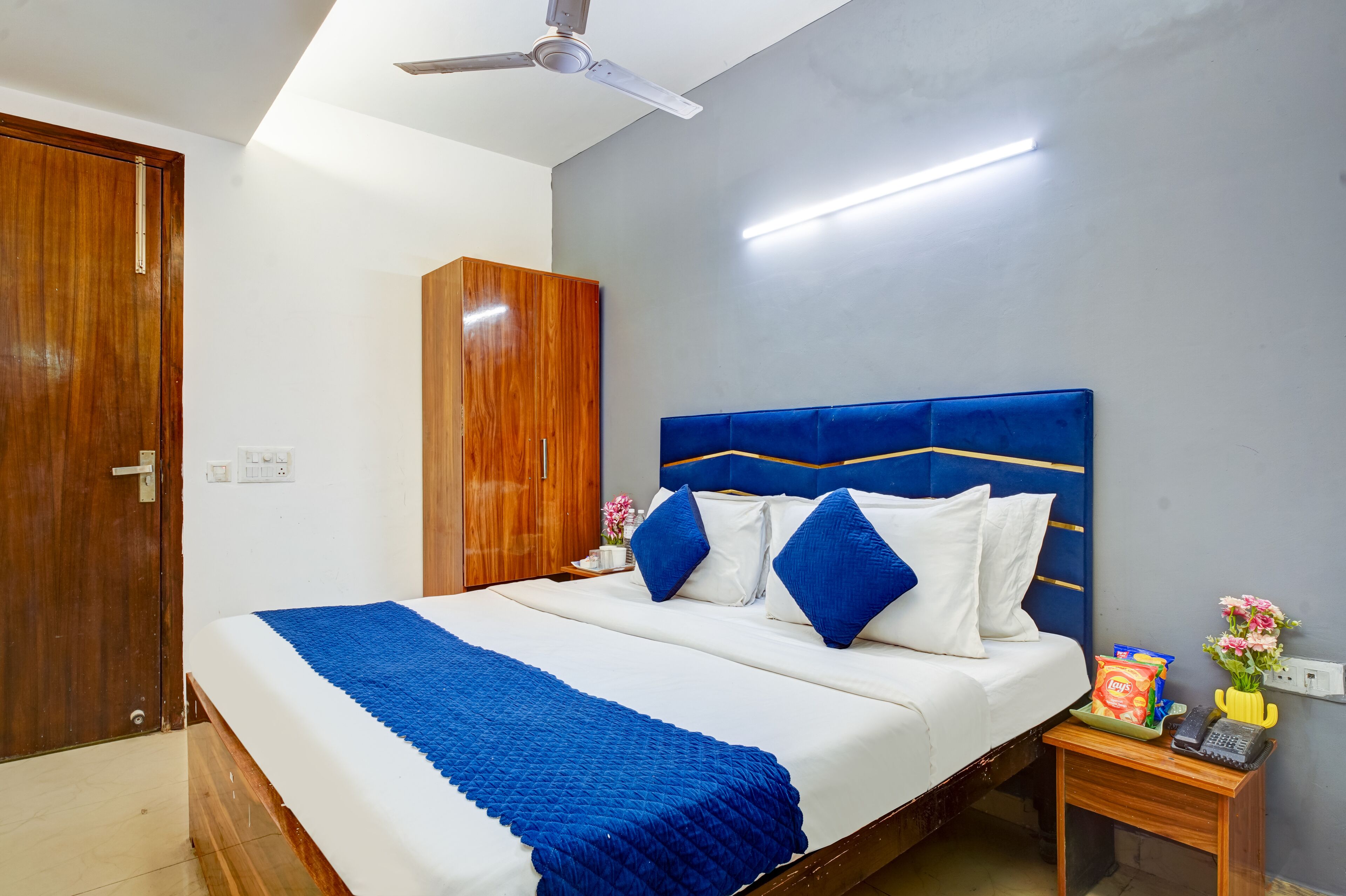 A neatly made bed with a blue blanket, a wooden wardrobe, a bedside table with a vase of flowers, and a ceiling fan.
