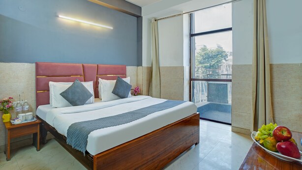 A hotel room with a large bed, a bedside table with a vase and cups, and a view of the city through a large window. - Hotel Black Rose (New Delhi)