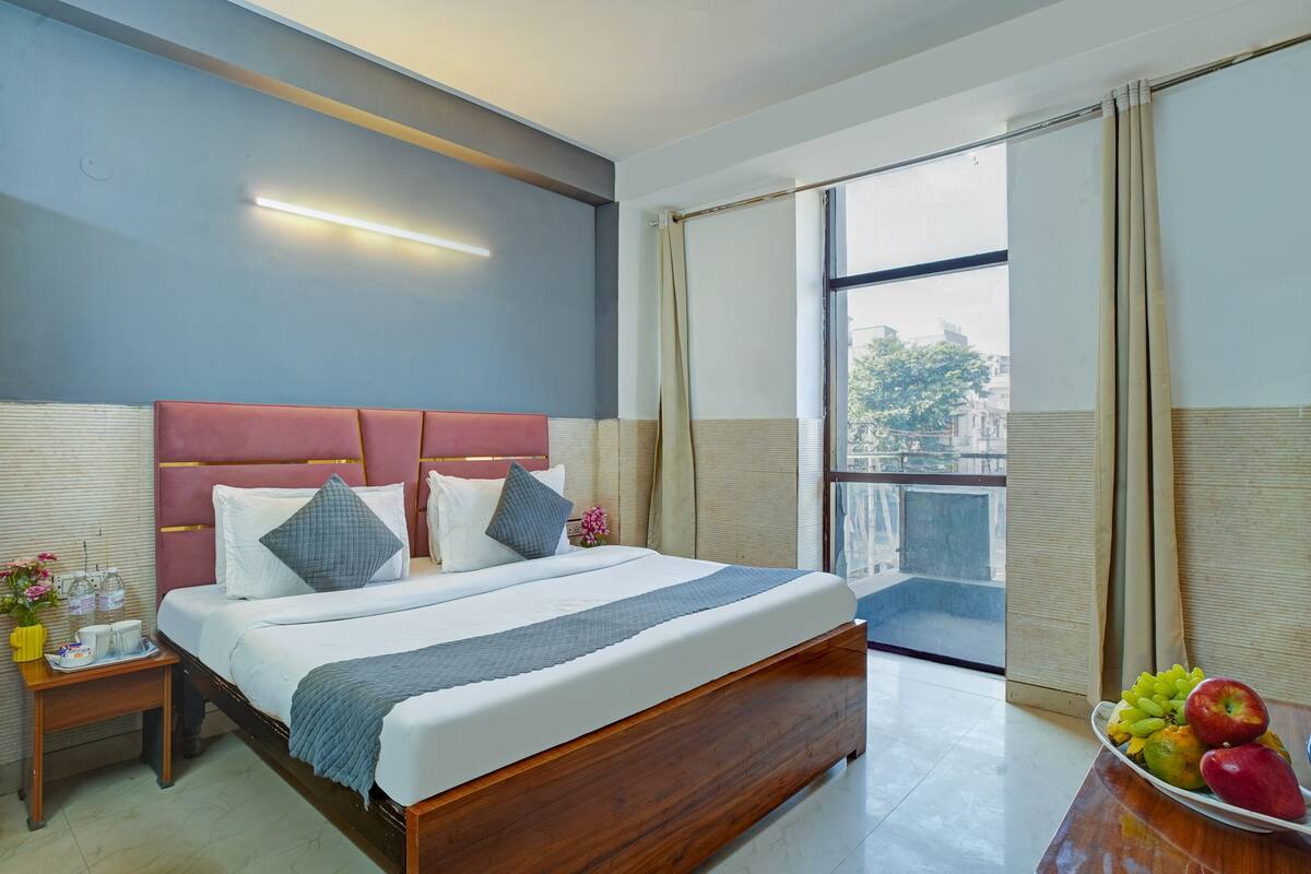 A hotel room with a large bed, a bedside table with a vase and cups, and a view of the city through a large window.