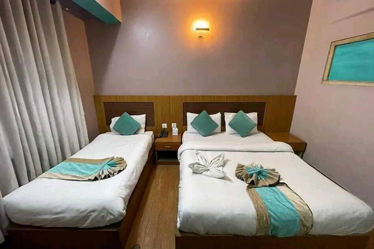 A hotel room with two beds, each with white linens and teal pillows. There is a nightstand with a phone and a small table between the beds. A wall-mounted light is above the beds, and a window with a view is on the right wall.