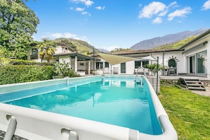 A rectangular swimming pool with a white deck, surrounded by lush greenery and a mountain backdrop.