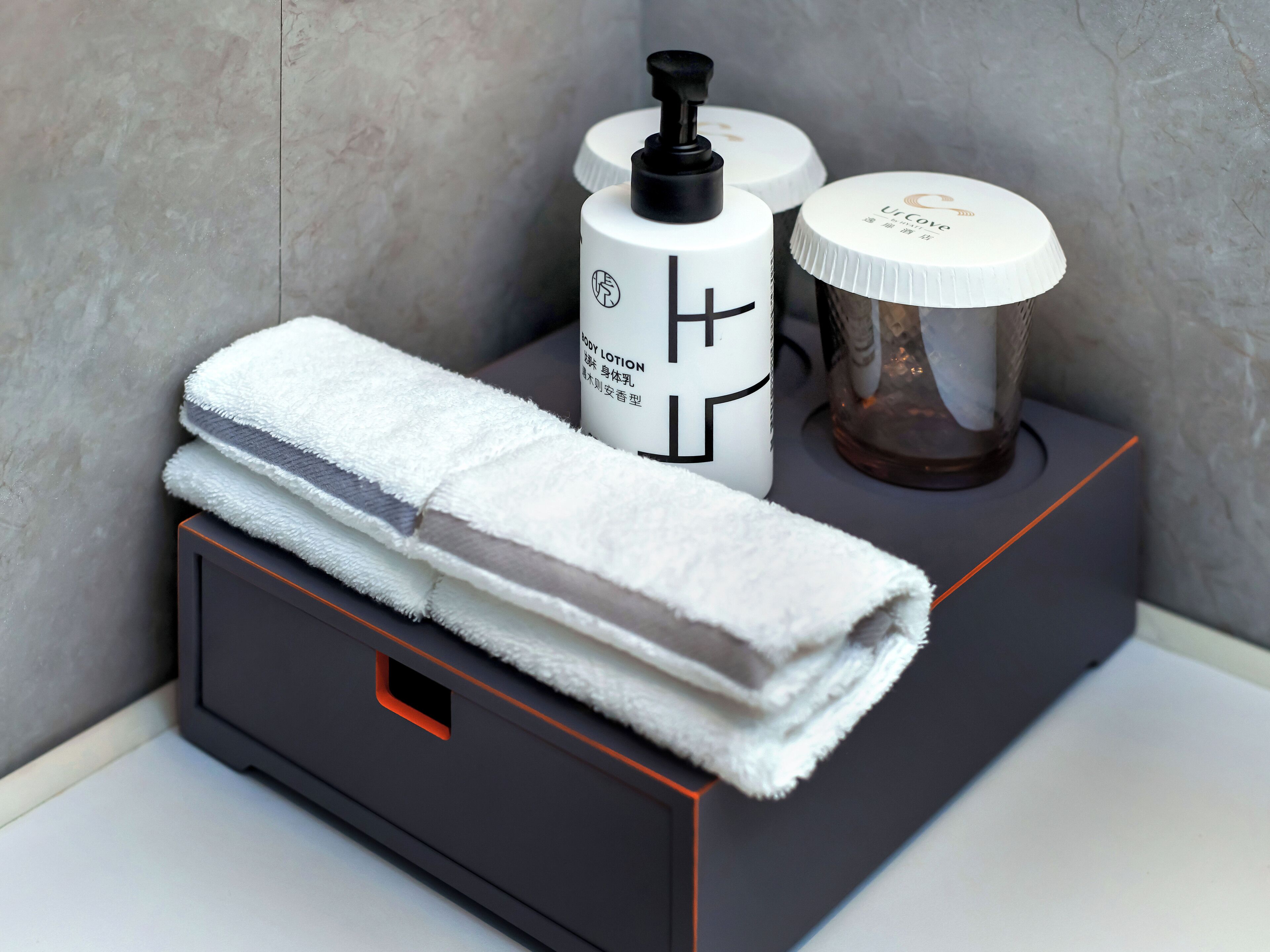 Een badkamer met een handdoek, een fles lotion en een glazen bakje op een zwarte doos.