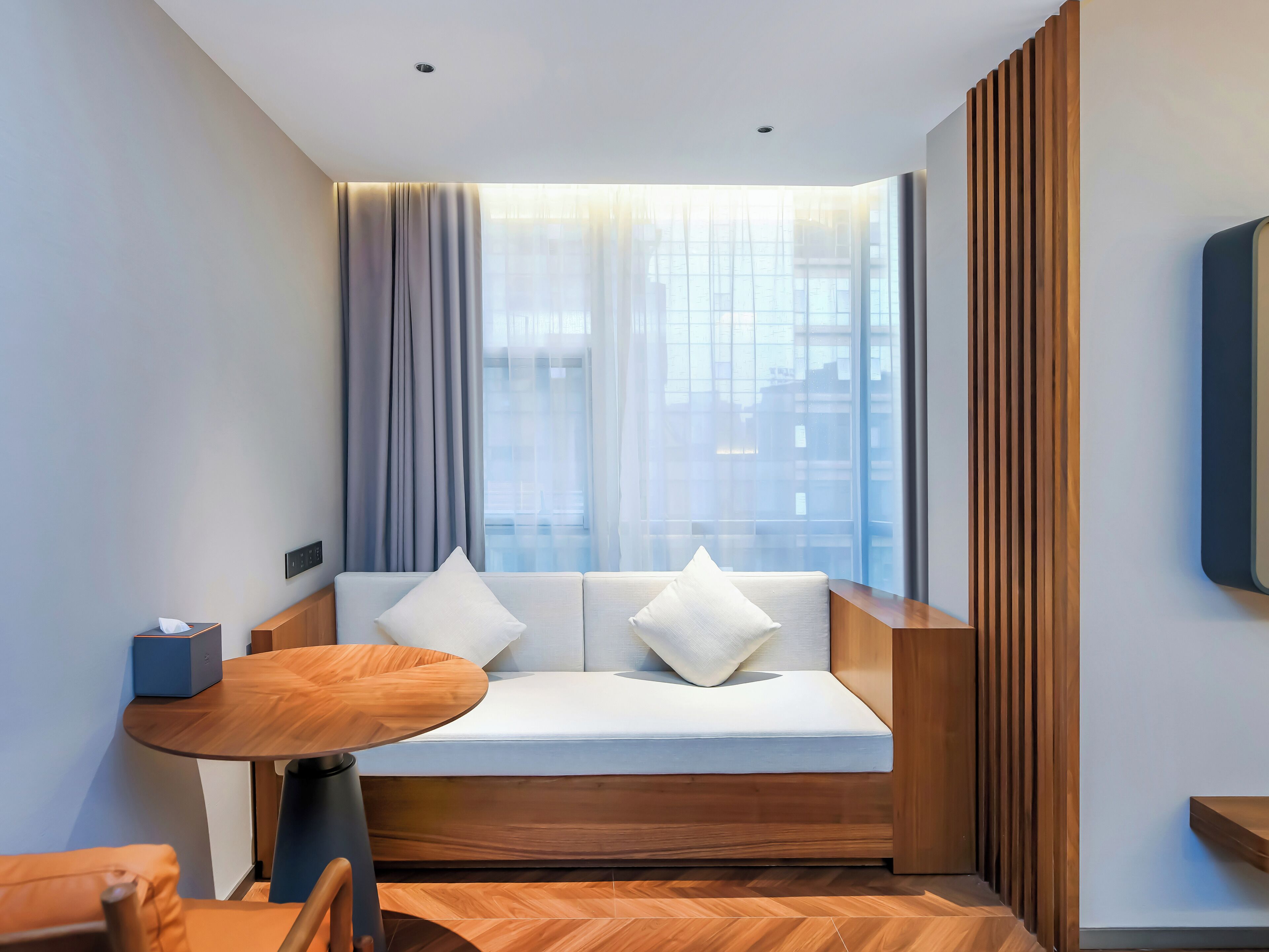 Habitación de hotel moderna con sofá, una mesita redonda y vistas a la ciudad a través de amplios ventanales.