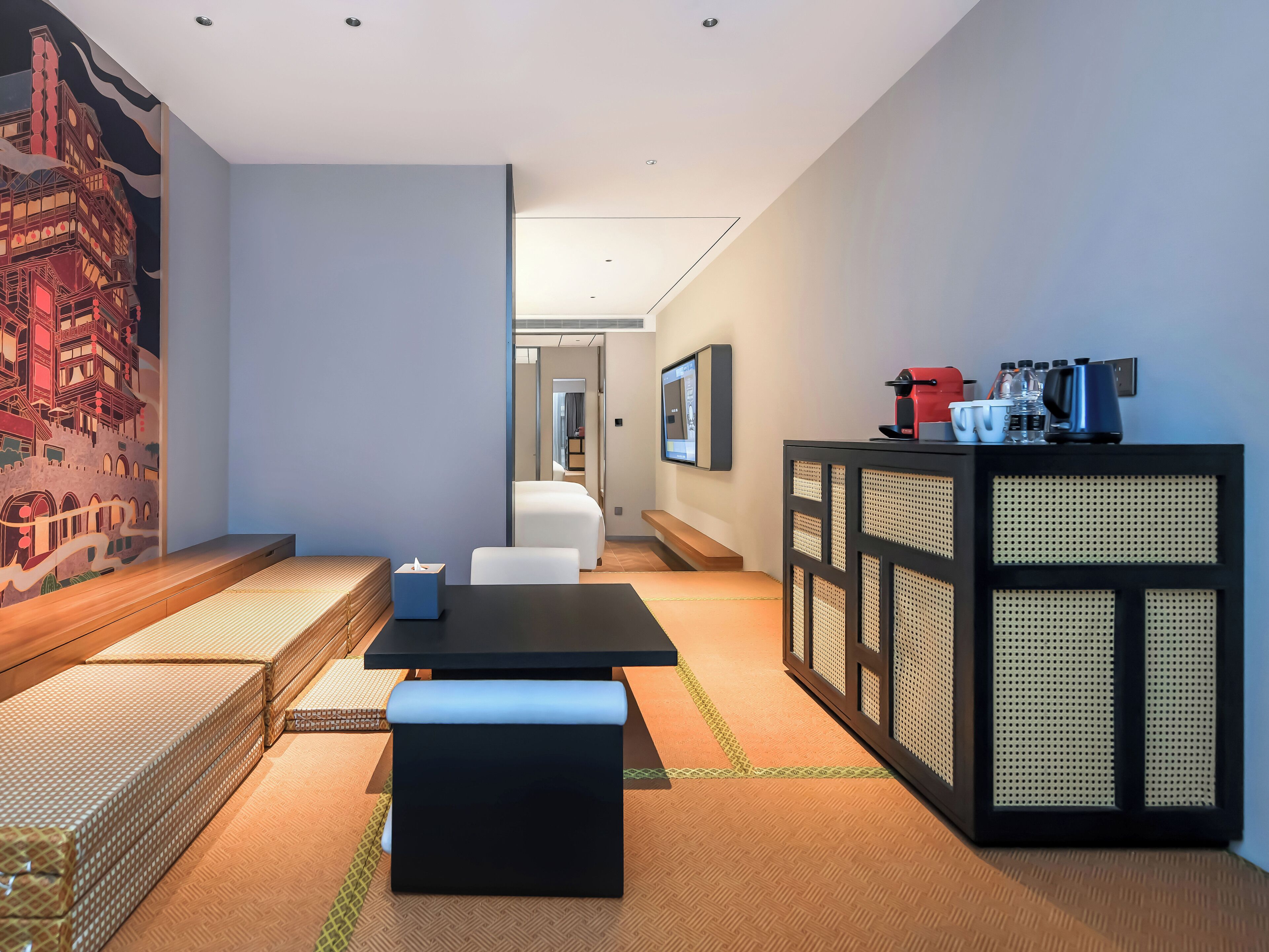 A modern hotel room with a sofa, a table, a bench, a wall-mounted TV, and a cabinet with kitchen items.