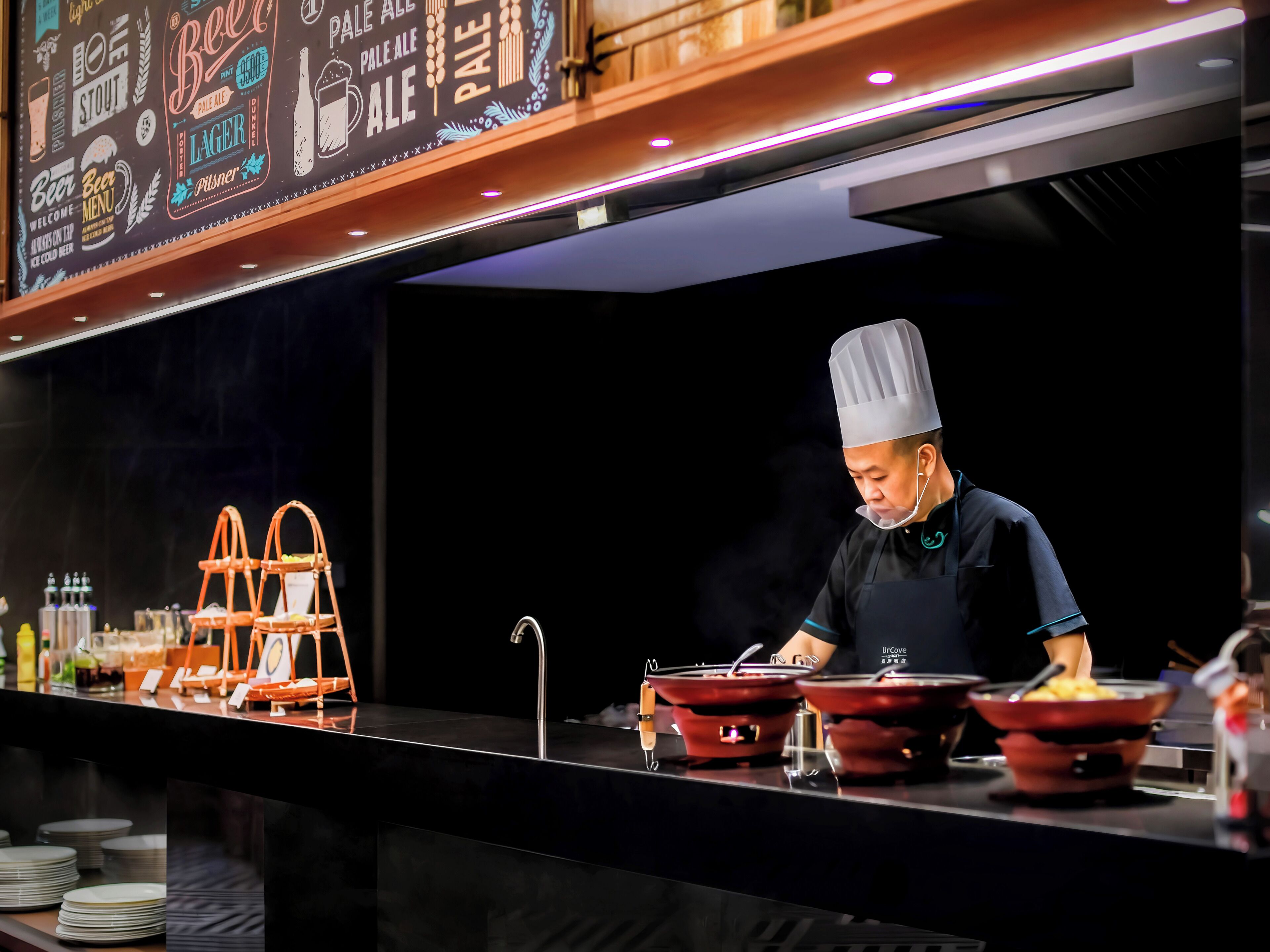 A chef in a professional kitchen with stainless steel counters and a menu board above.