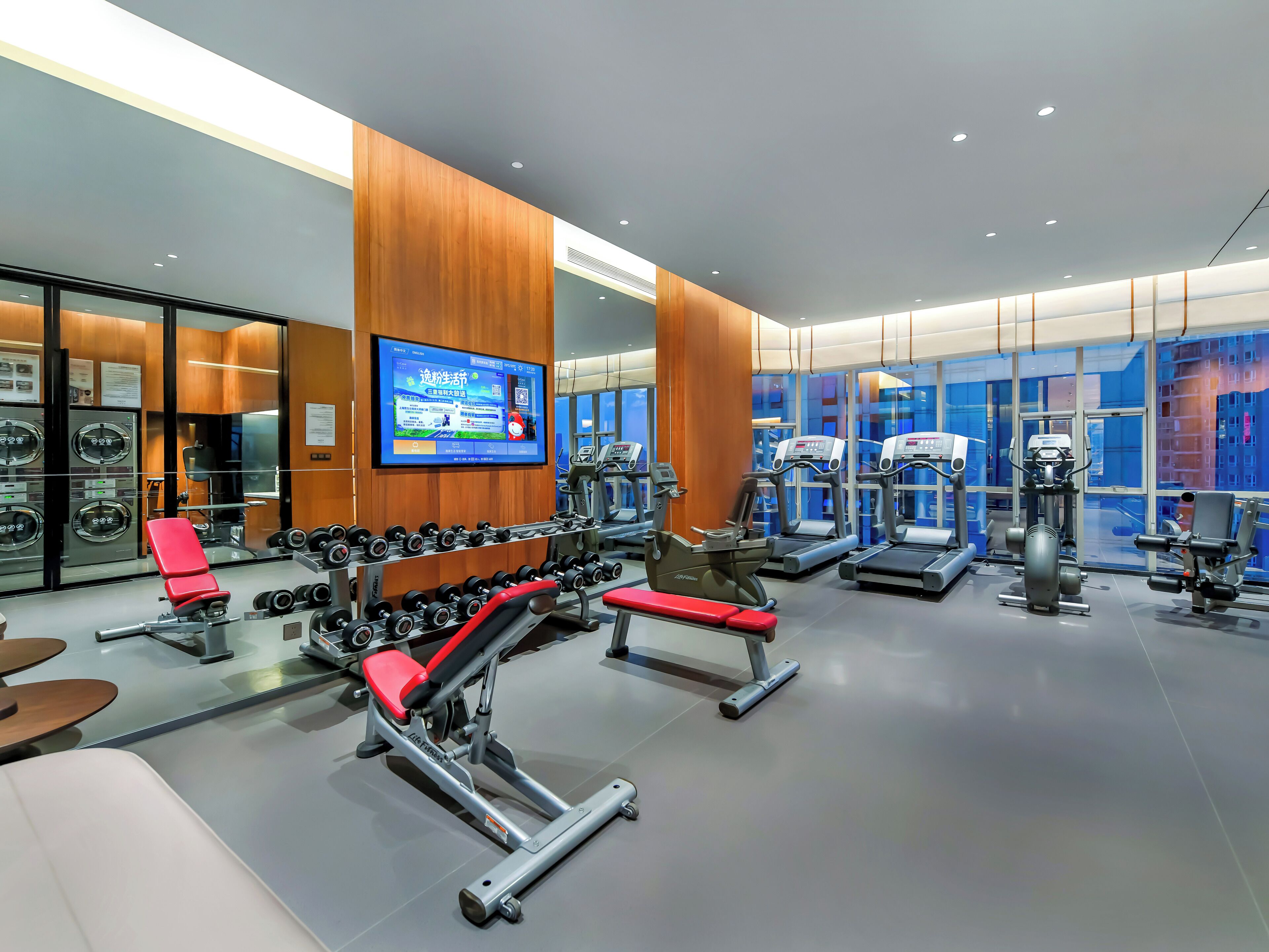 A modern gym with various exercise equipment, a large mirror, and a digital screen displaying workout information.