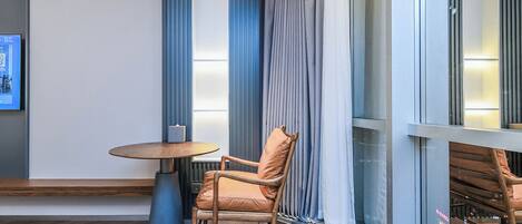 A modern hotel room with a wooden floor, a round table, a cushioned chair, and a large window with curtains.