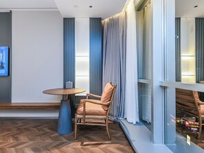 Une chambre d’hôtel moderne avec un plancher en bois, une table ronde, un fauteuil rembourré et une grande fenêtre avec des rideaux.