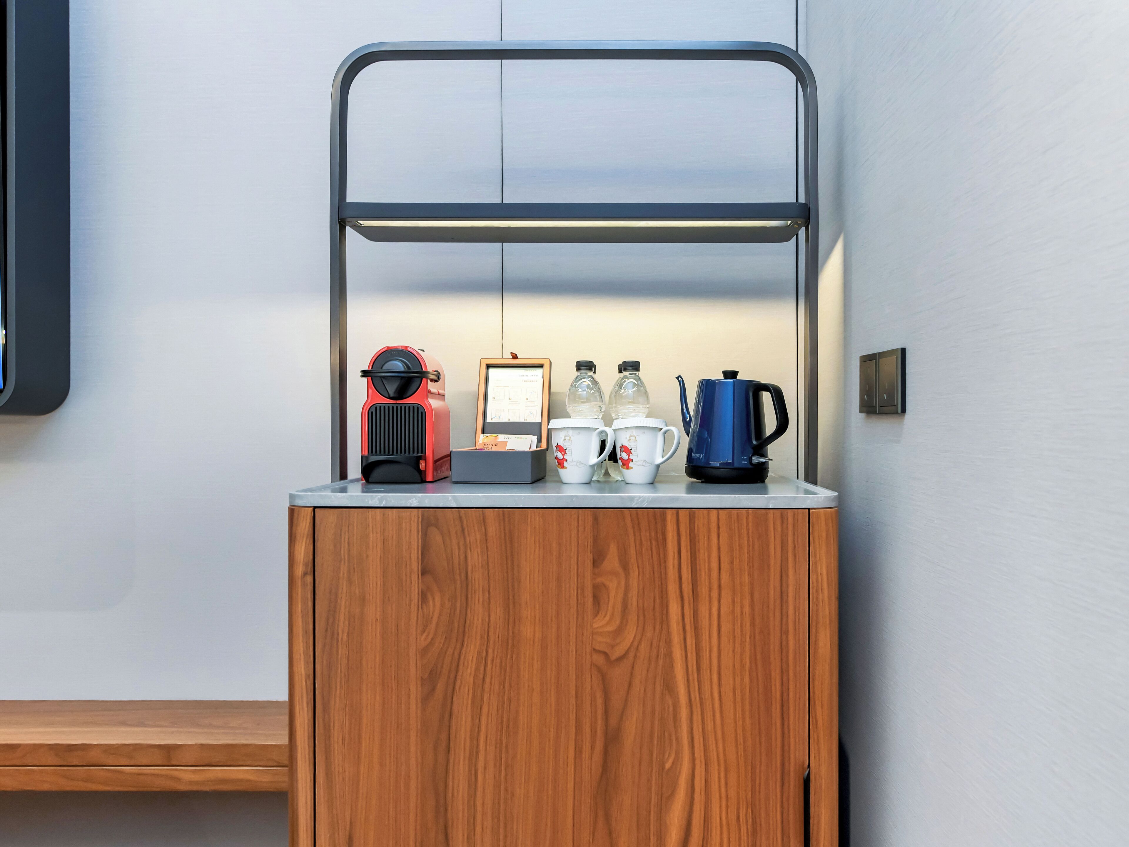 A wooden cabinet with a metal shelf containing a red appliance, a framed document, two water bottles, and a blue kettle.