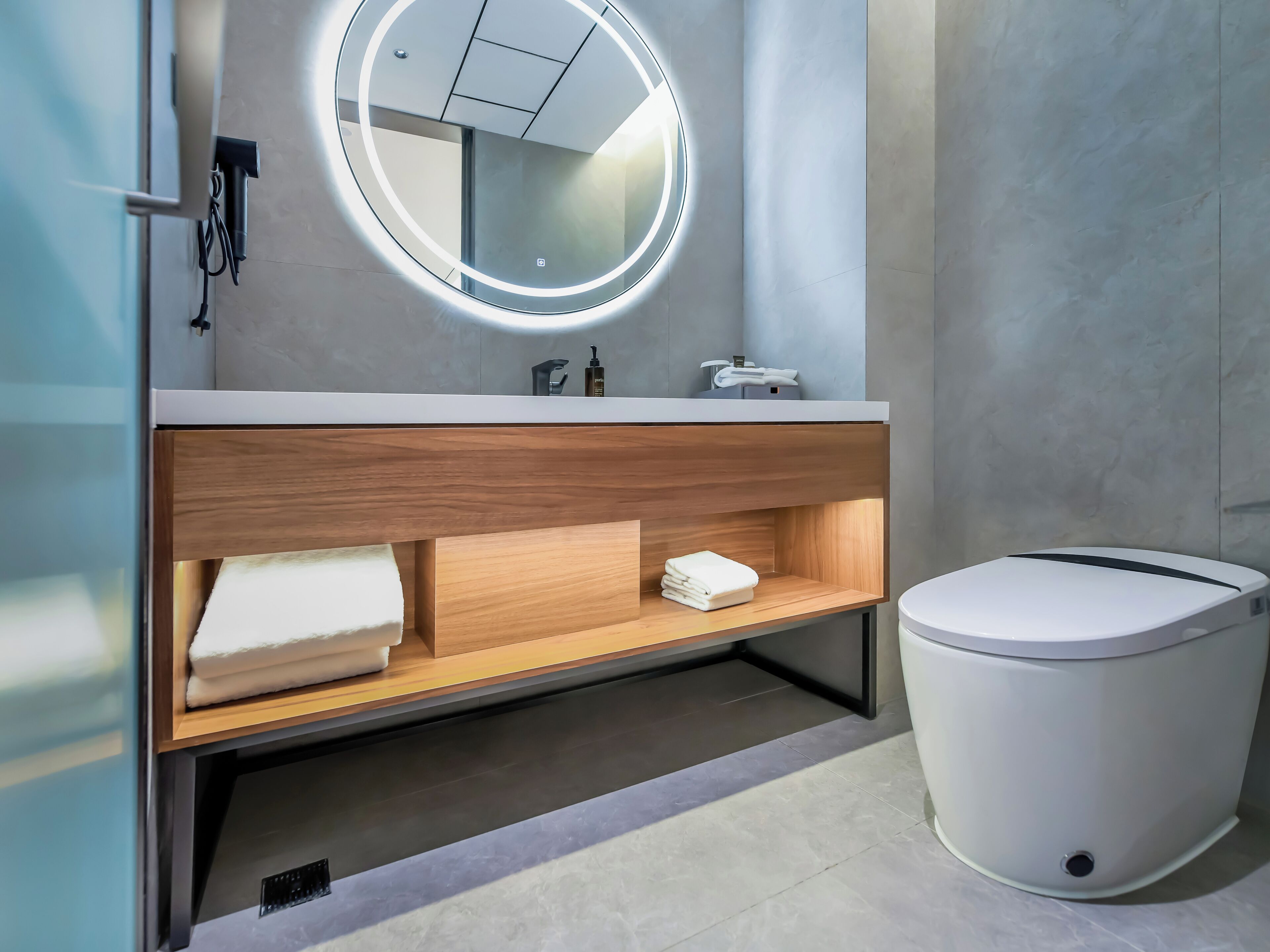 A modern bathroom with a round mirror, wooden vanity, white toilet, and a towel rack.