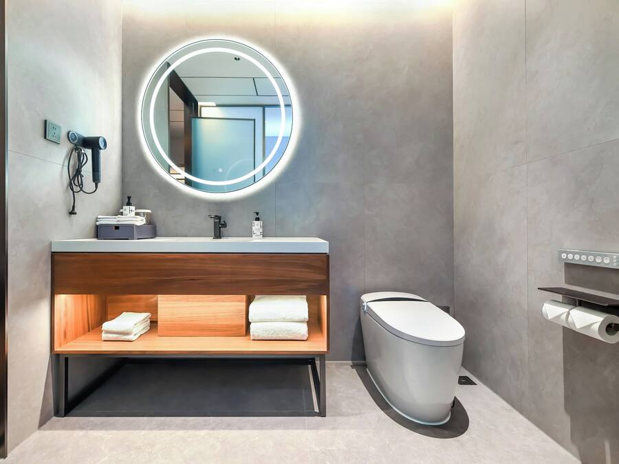 A modern bathroom with a round mirror, a sink, a toilet, and a wooden shelf with towels.