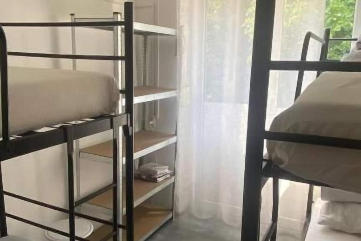 A dormitory room with bunk beds, a window with sheer curtains, and a view of greenery outside.