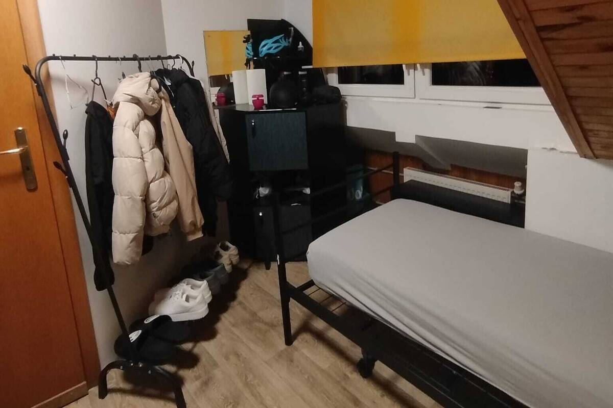 A small, compact room with a bunk bed, a desk with a chair, a clothing rack, and a window with blinds.