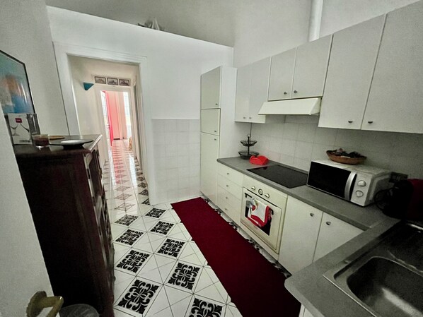 A compact kitchen with white cabinets, a microwave, an oven, and a sink. A red rug is on the floor, and the walls are tiled. - 75m2 apartment for 4 pax a few steps from Piazza Tasso (Sorrento)