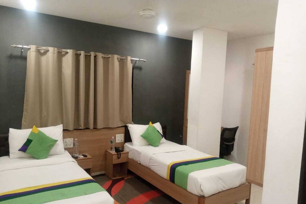 A hotel room with two single beds, a nightstand with a lamp, a small table with a bottle and a remote control, and a large window with curtains.