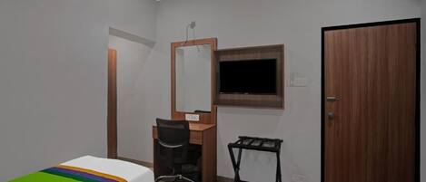 A small, well-lit room with a wooden desk, a chair, a bed with a colorful blanket, a television mounted on the wall, and a door.