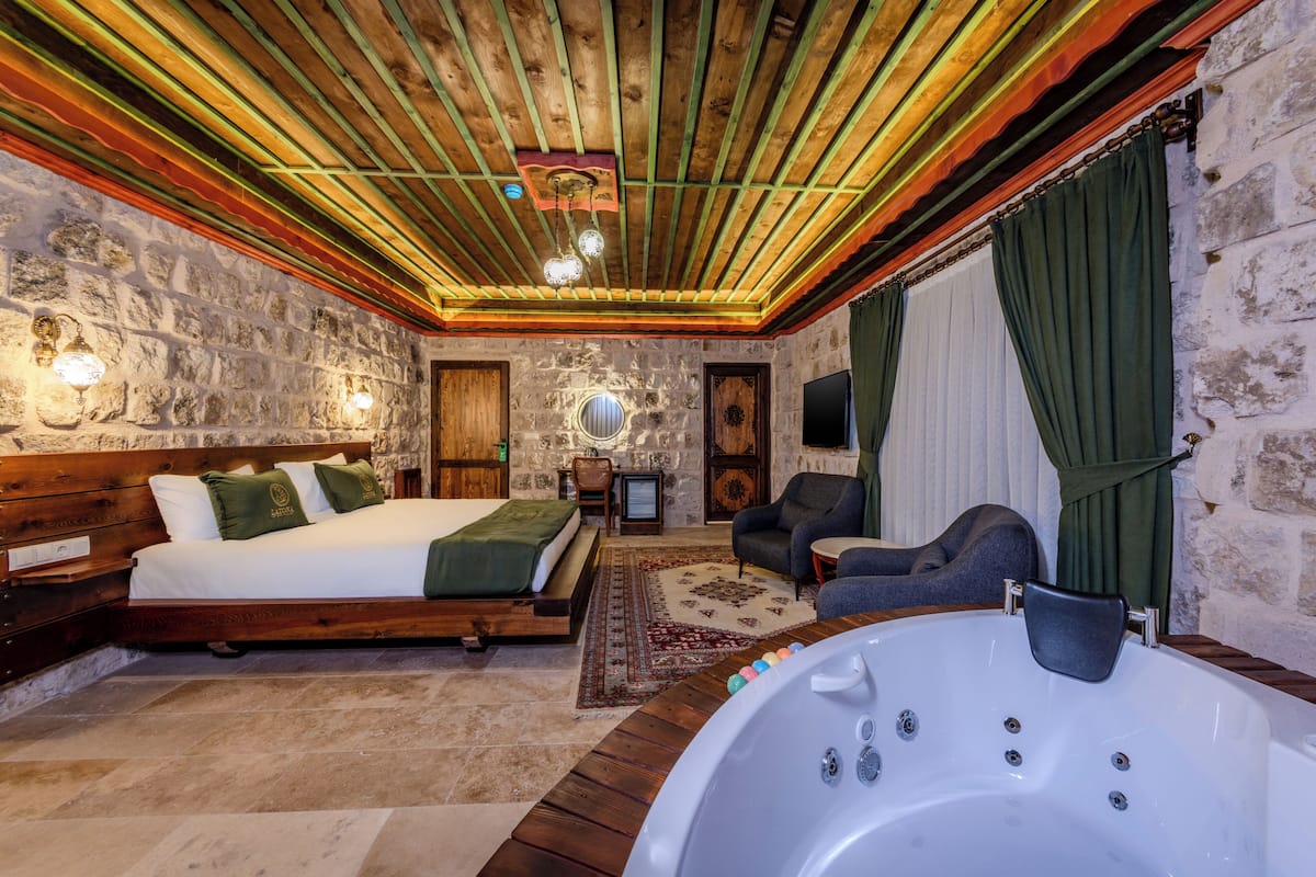 Una habitación con techo de madera, paredes de piedra, una cama con ropa de cama verde, un rincón de descanso con una silla y una mesa, y una bañera grande.