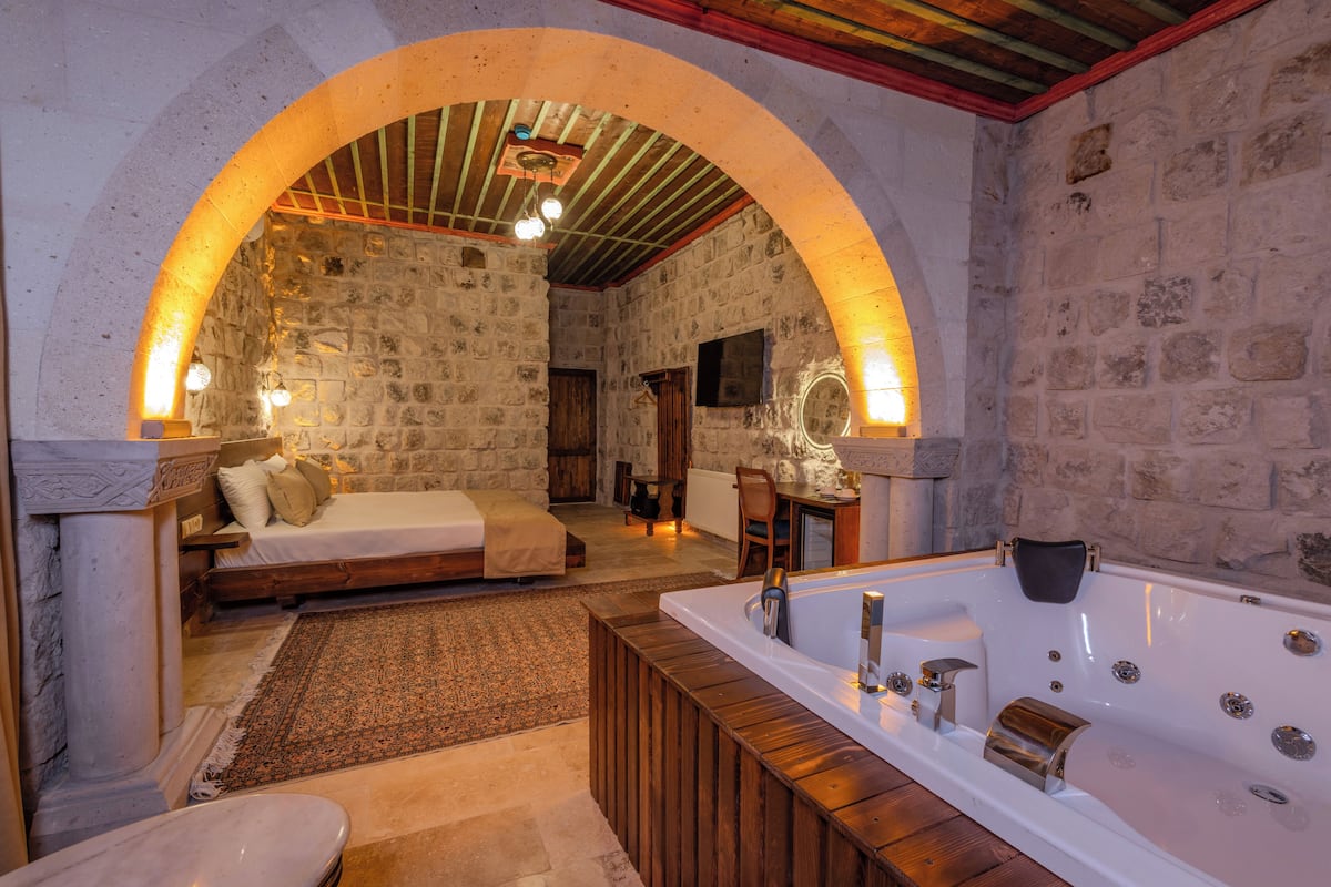 Una habitación con paredes de piedra, un banco de madera, una bañera grande y una cama con cabecero de madera.