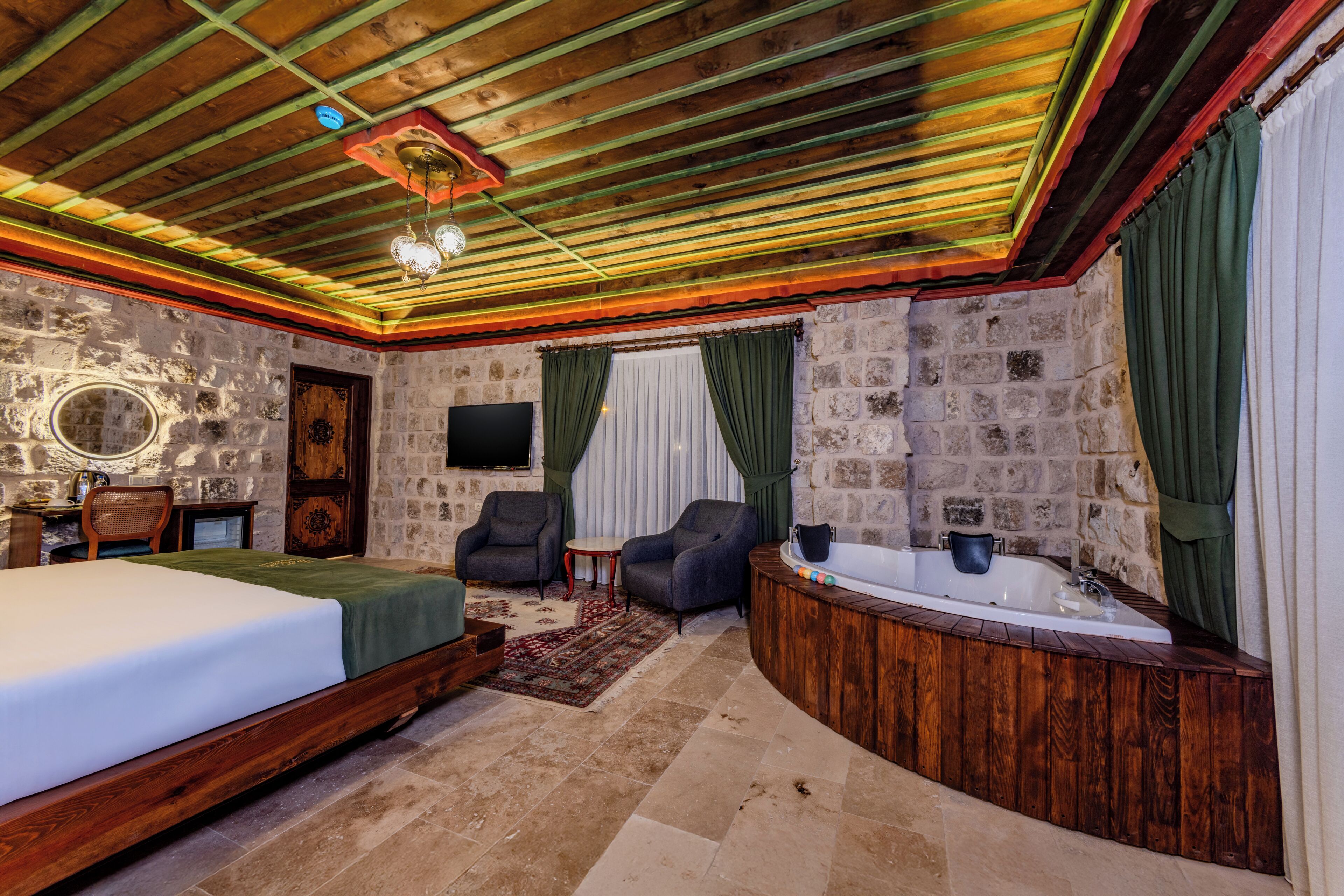 A room with a bed, two armchairs, a small table, a TV, a bathtub, and stone walls.