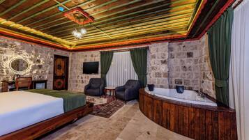 A room with a bed, two armchairs, a small table, a TV, a bathtub, and stone walls.