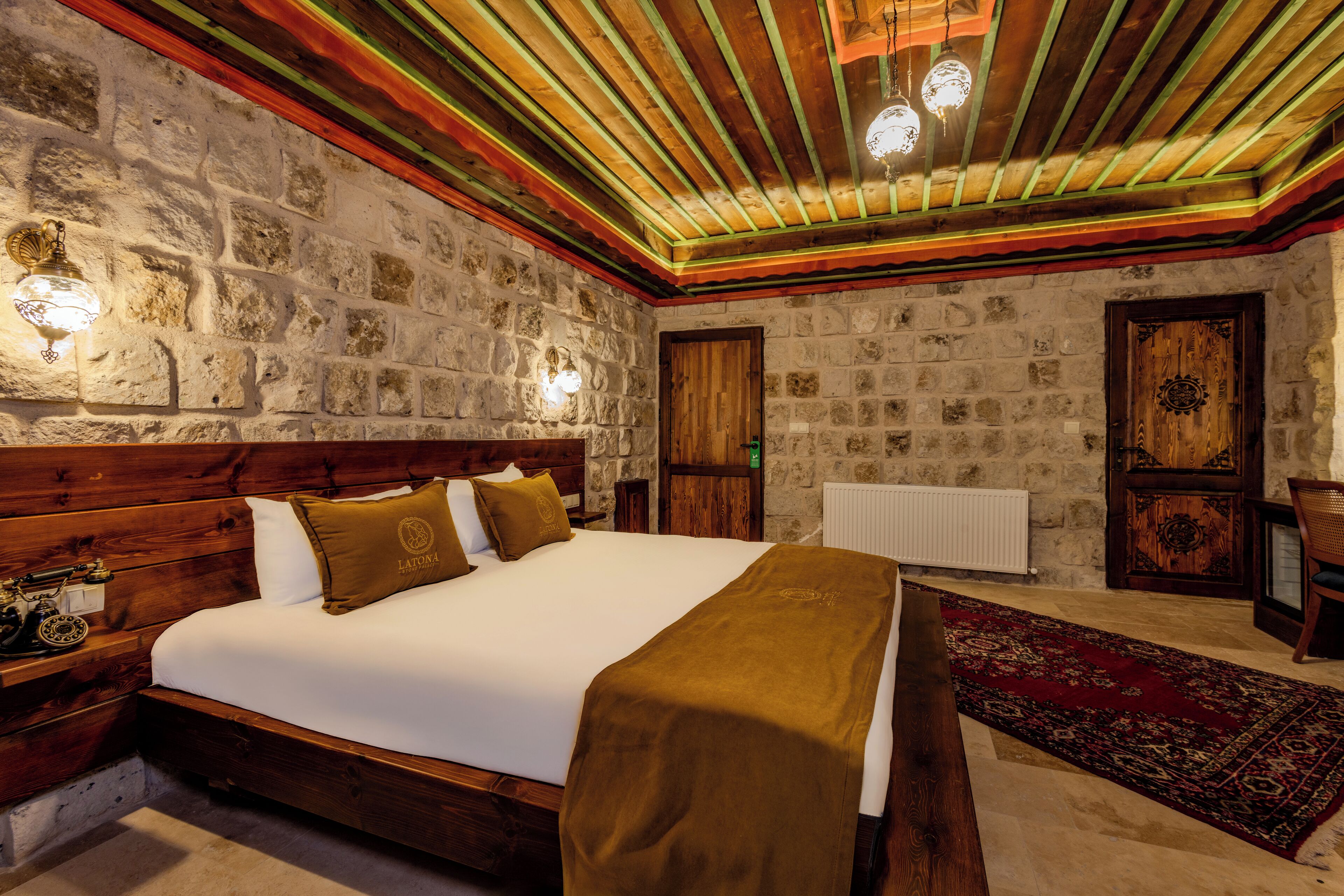 Une chambre dotée d’un grand lit, de murs en pierre, de poutres en bois au plafond et d’une moquette rouge.