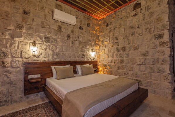 Un dormitorio con paredes de piedra, una cama de madera, dos mesitas de noche e iluminación fija en la pared.