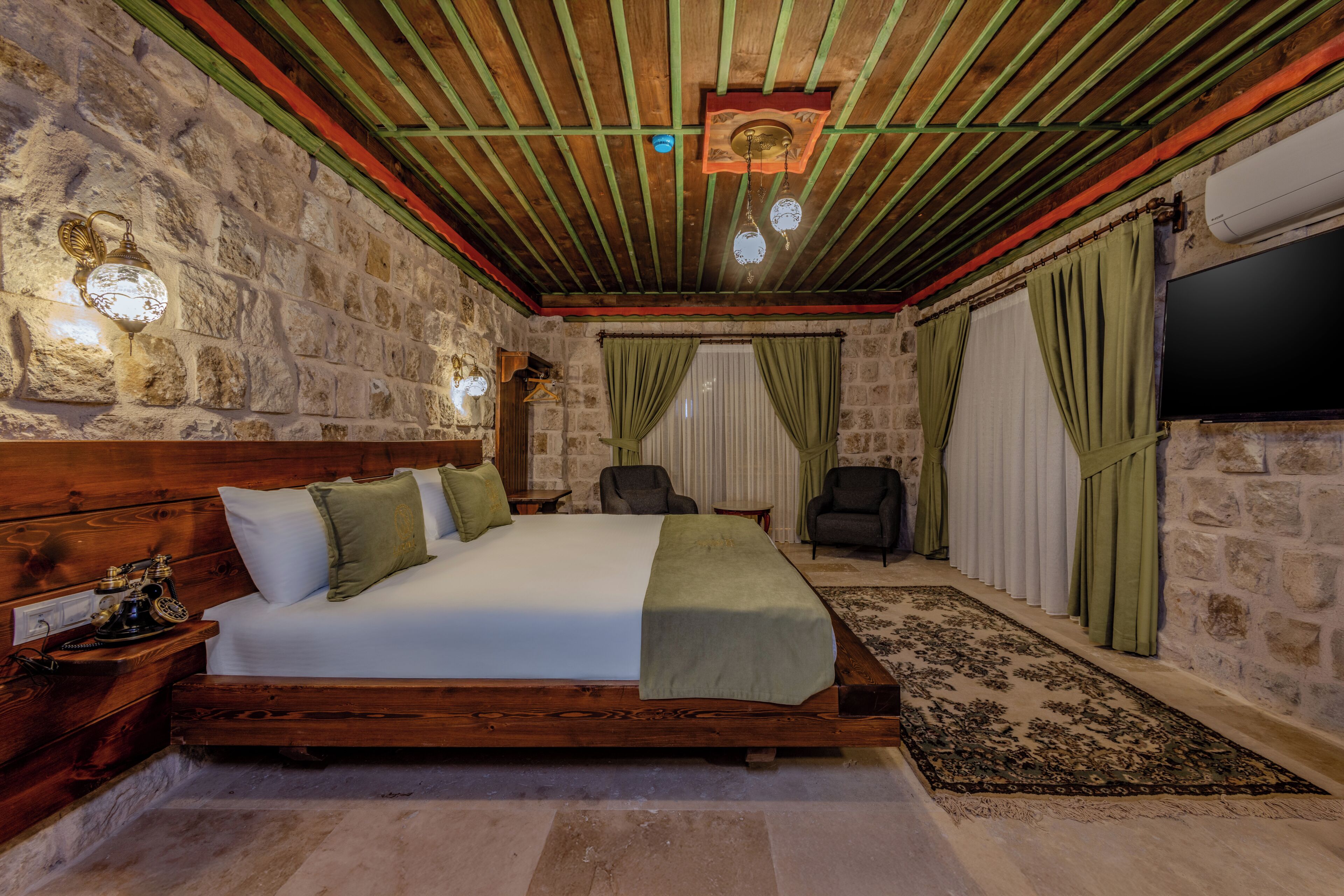 A room with a stone wall, a wooden bed, green curtains, a flat-screen TV, and a stone floor.