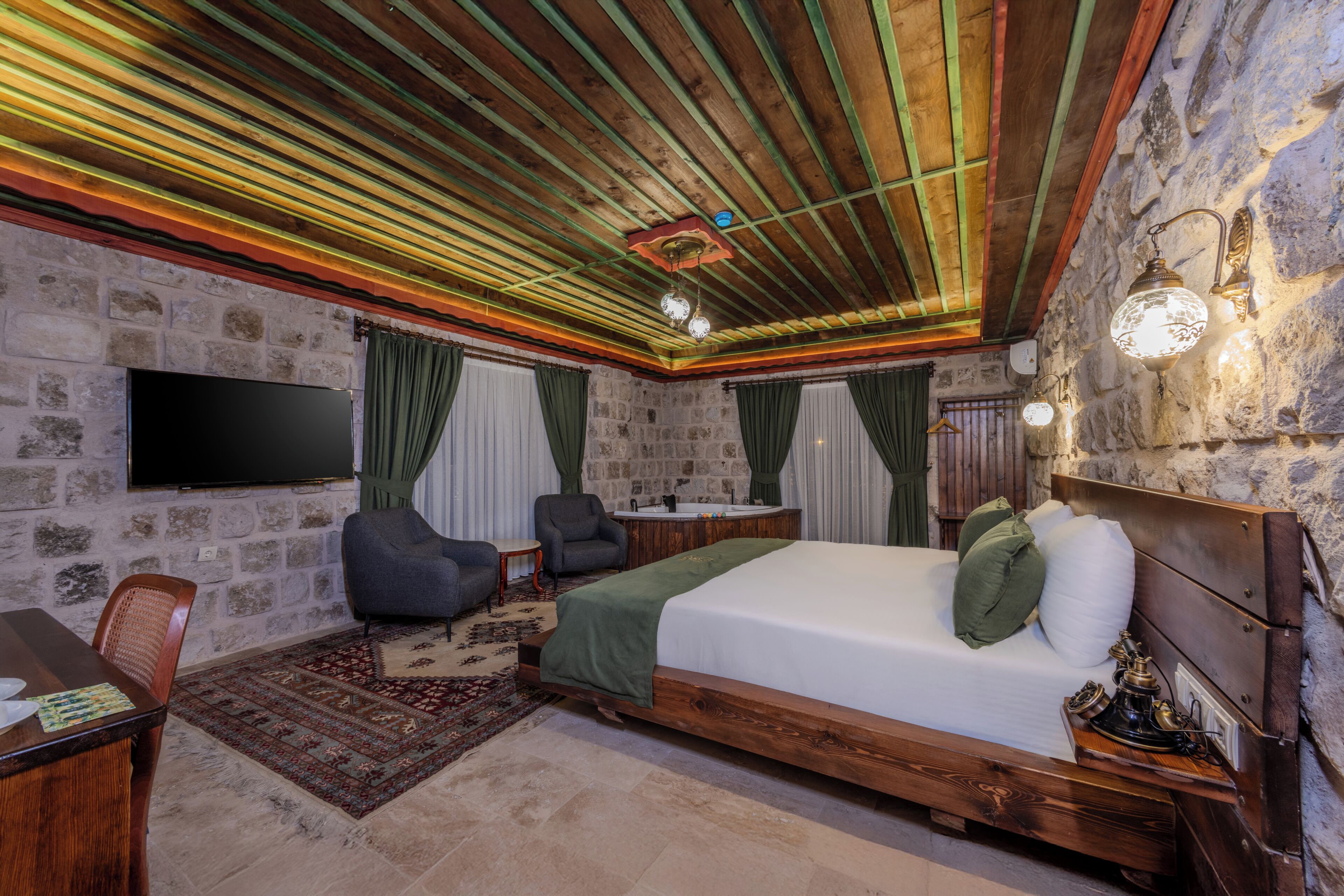 A bedroom with a bed, a TV, a desk, a chair, and a stone wall.