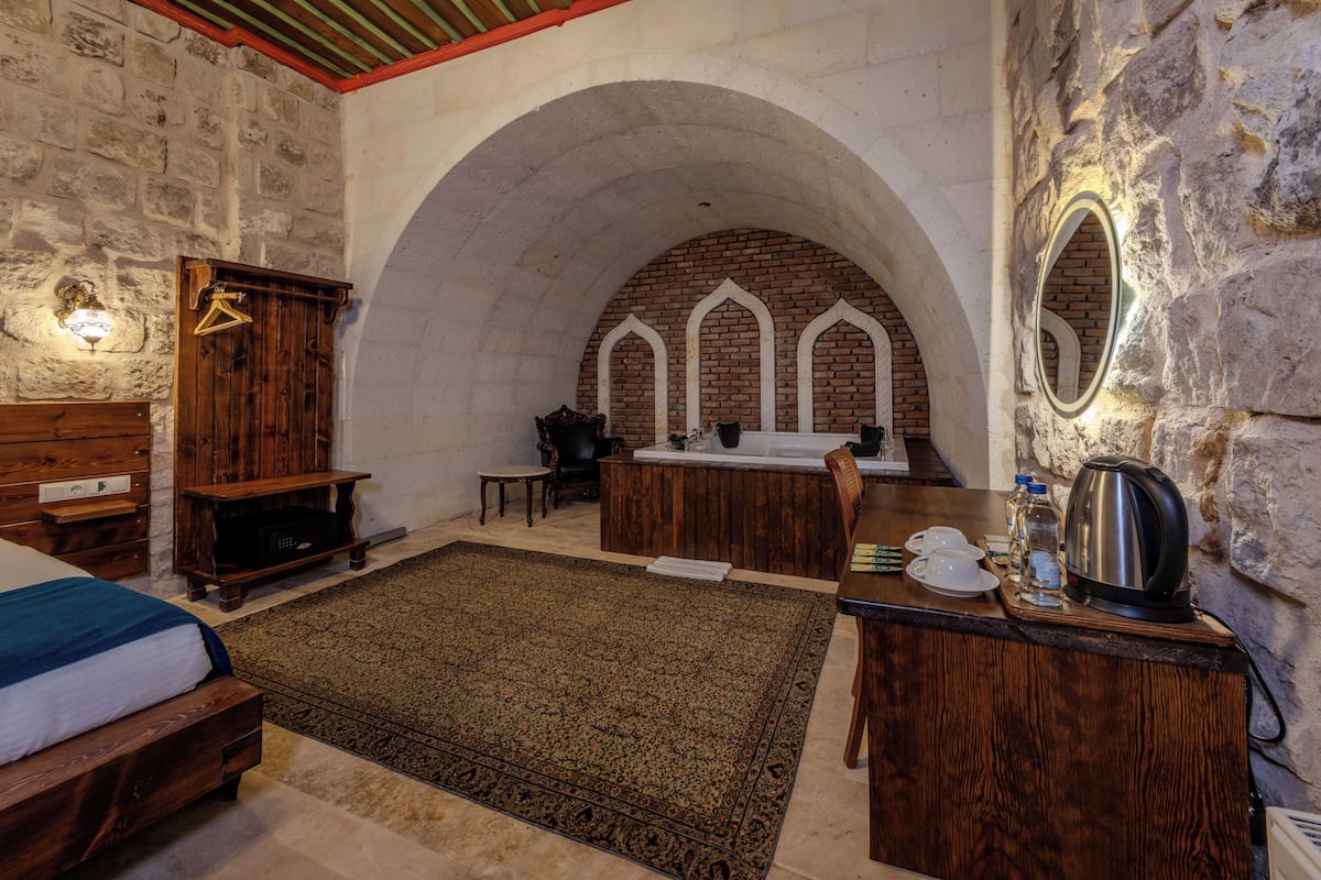 Una habitación con cama, muebles de madera, un escritorio con hervidor y una pared de piedra con nichos arqueados.