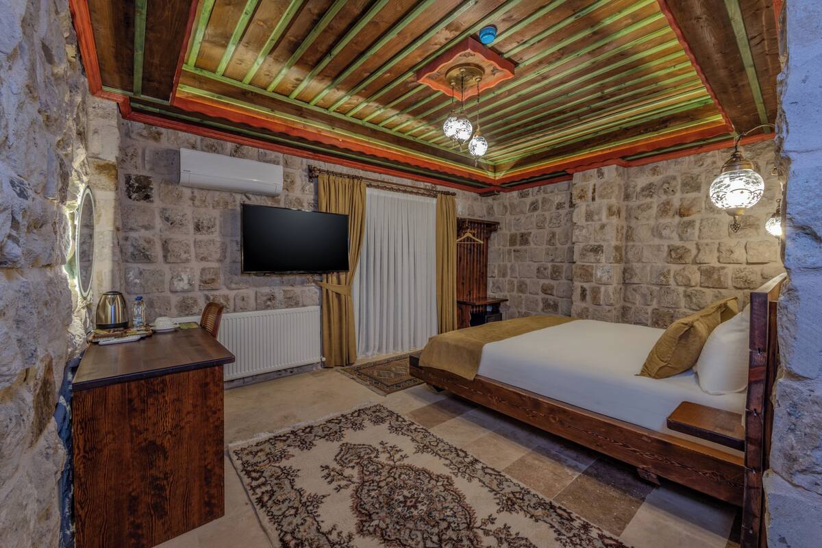 Un dormitorio con pared de piedra, techo de madera, una cama, un televisor y un escritorio.