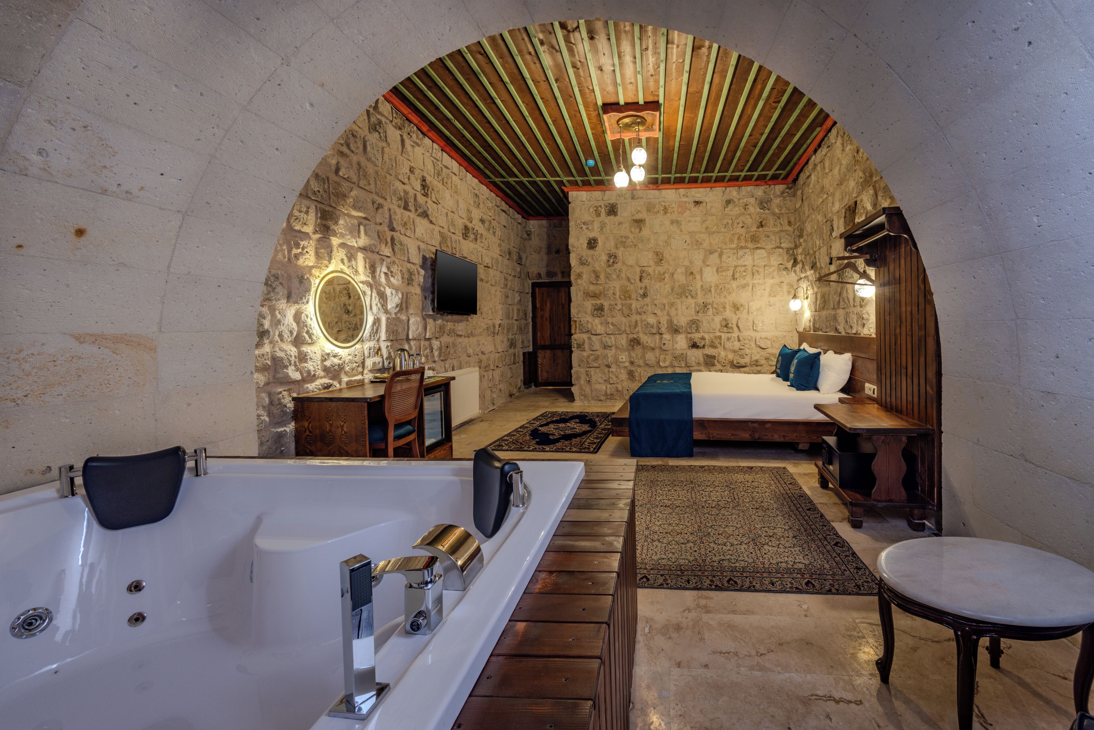 A room with a stone wall, wooden ceiling, a large bathtub, a bed with blue bedding, a bedside table, and a small table with a chair.