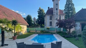 Ein Schwimmbecken mit Liegestühlen, eine Steinmauer und ein großes Gebäude mit einem Turm im Hintergrund.