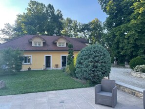 Une maison à deux étages avec une façade jaune, un toit brun et un jardin devant avec une pelouse et un grand conifère.