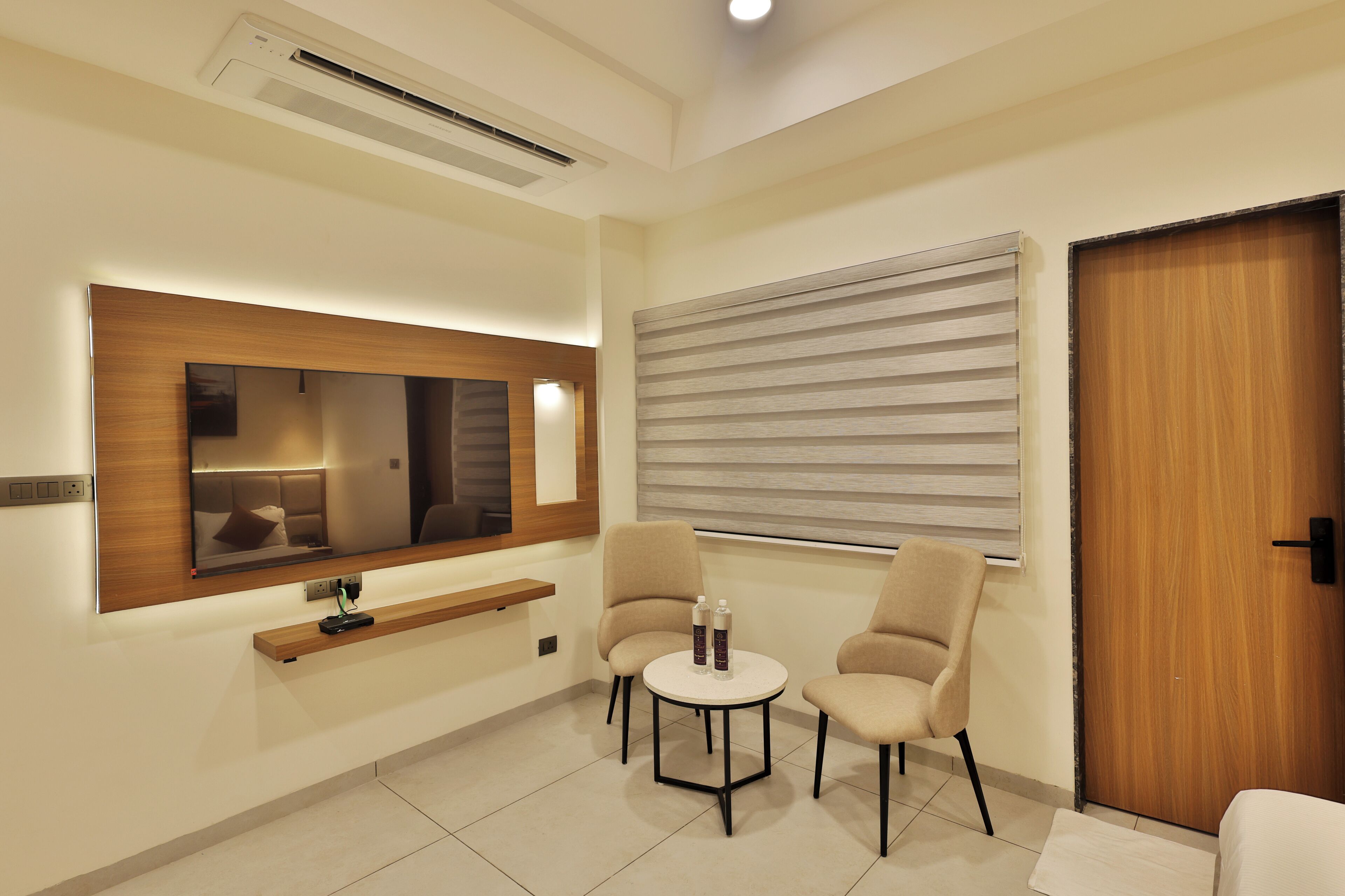 A modern hotel room with a large mirror, two chairs, a small table, and a wooden door.