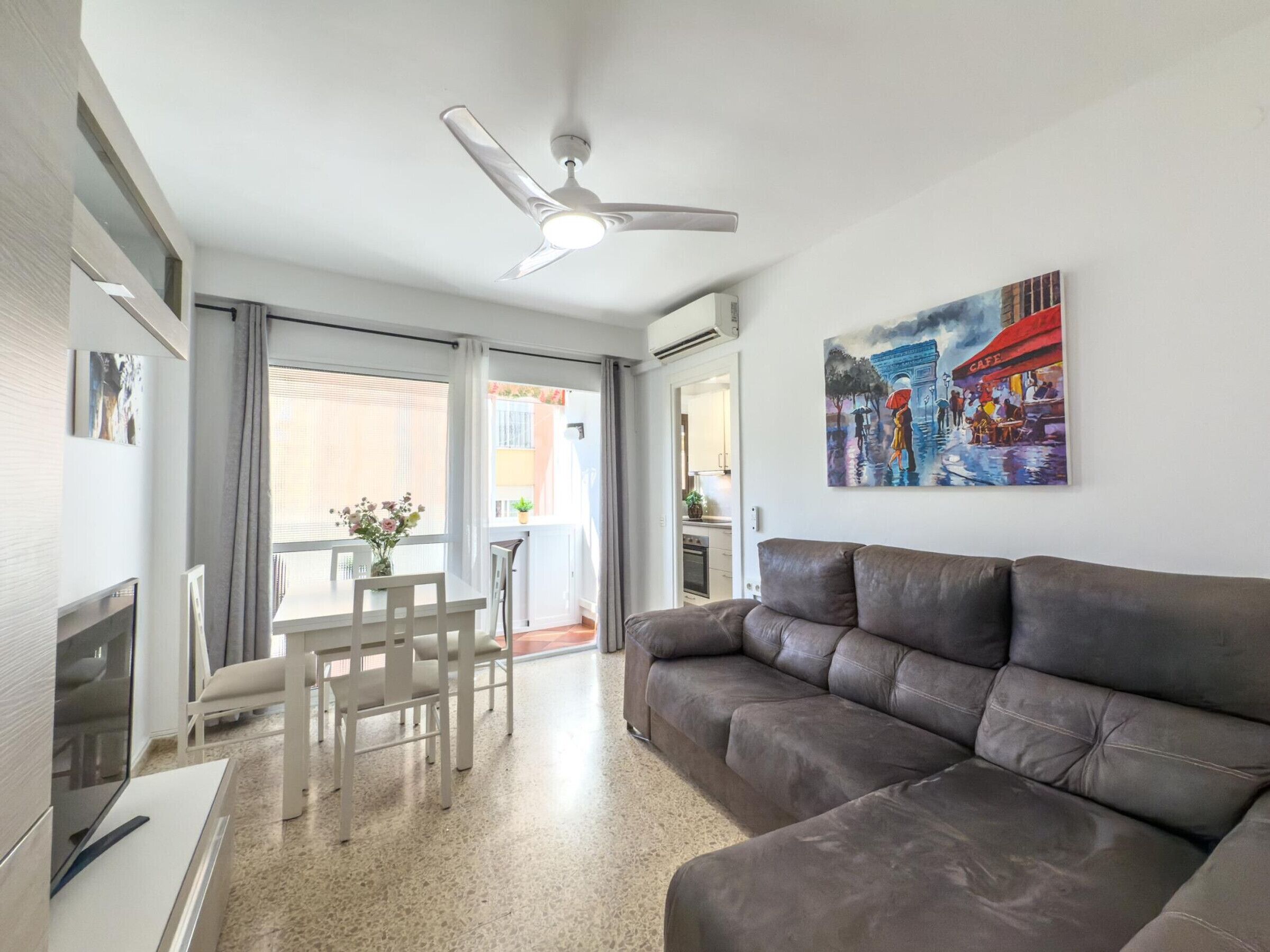 A modern living room with a leather sofa, a dining table with chairs, a ceiling fan, and a painting on the wall.