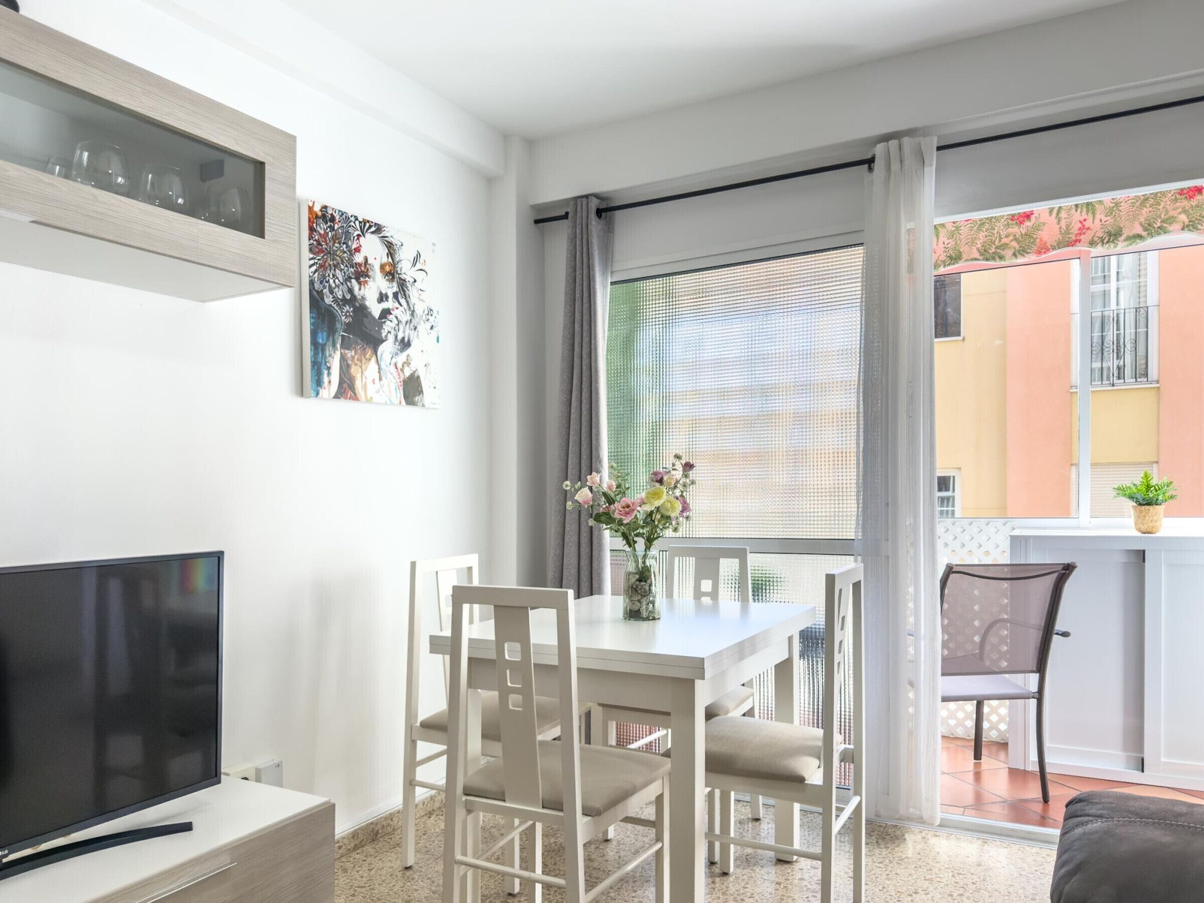Ein modernes Wohnzimmer mit Flachbildfernseher, einem Esstisch mit Stühlen, einer Vase mit Blumen und einem Balkon mit einem Blumentopf.