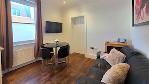 Een moderne woonkamer met een zwarte leren bank, een ronde eettafel met twee stoelen, een flatscreen televisie aan de muur en een houten bijzettafeltje met een klok.