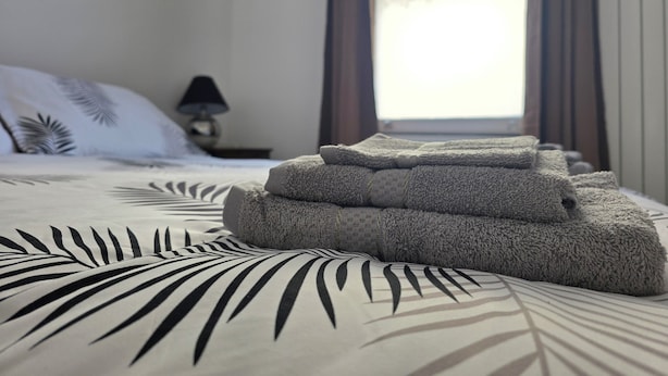 Een net opgemaakt bed met een bedsprei met een patroon, een stapel opgevouwen handdoeken, een bedlamp en een raam met gordijnen.