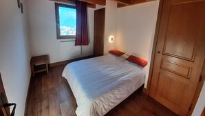 A bedroom with a wooden ceiling, a bed with white bedding and red pillows, a small wooden stool, a window with a view, and a wooden door.