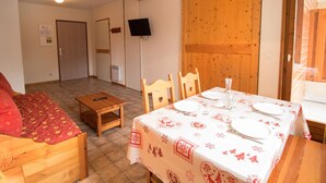 A cozy room with a wooden bed, a small table with a remote control, a dining table set with plates and cutlery, and a television mounted on the wall. - Cozy apartment with parking, sleeps 4, near shuttles in Aussois (Aussois)